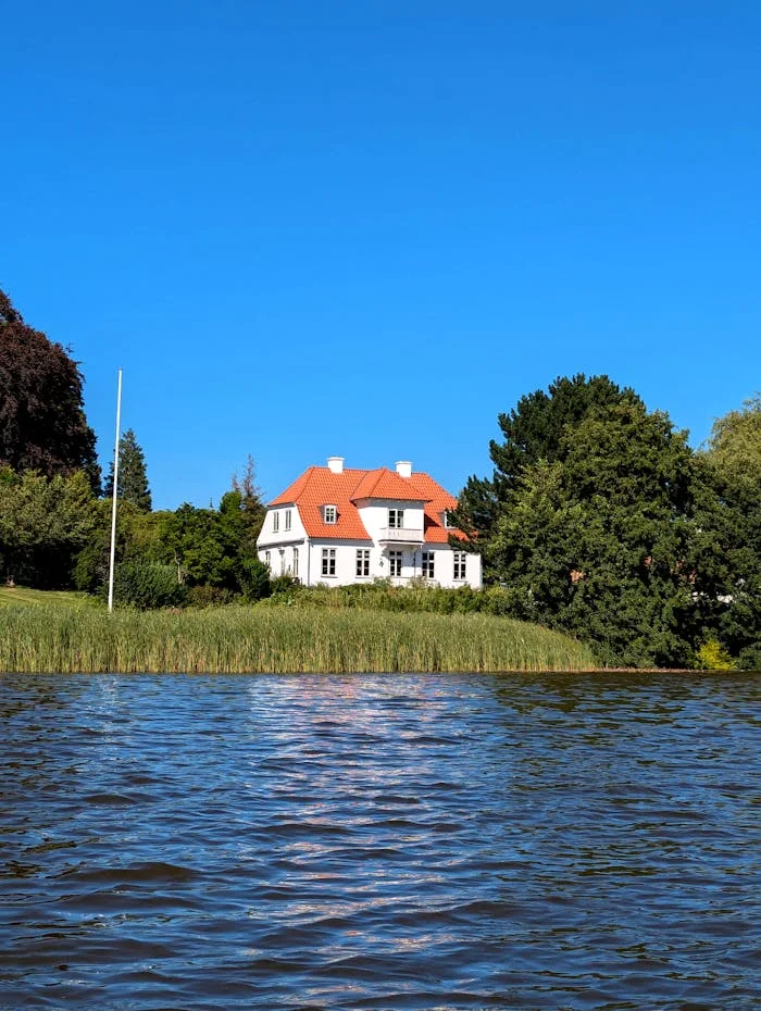 A picturesque white house with an orange roof by the lake under a clear blue sky.